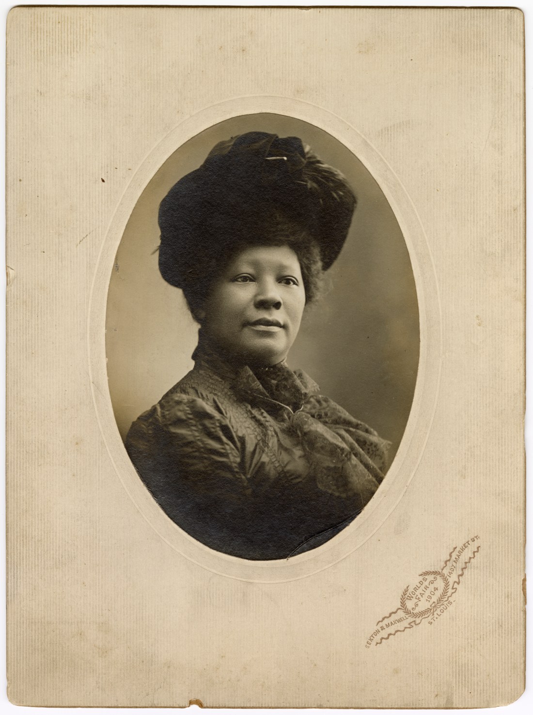 Studio portrait of an African American woman in 1904 wearing a large structured hat and lace bow, shown in three-quarter view on an oval photo mount stamped “World’s Fair 1904 / Sexton & Maxwell / 1407 Market St., St. Louis.”